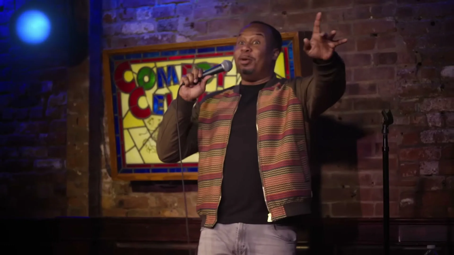 A Black male comedian in his 30s performs stand-up comedy on stage at a brick-walled club, holding a microphone and gesturing.