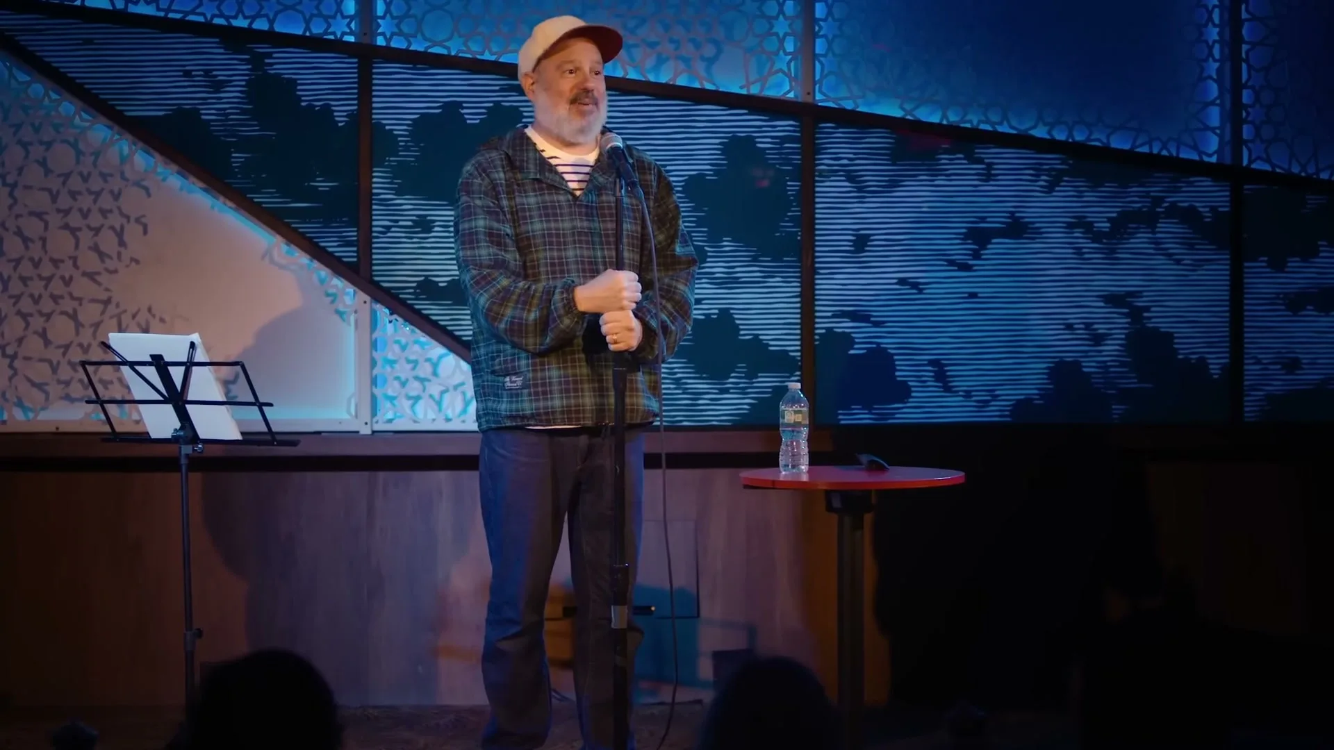 A male comedian with a beard and a cap stands on stage, holding a microphone and smiling at the audience in a comedy club.