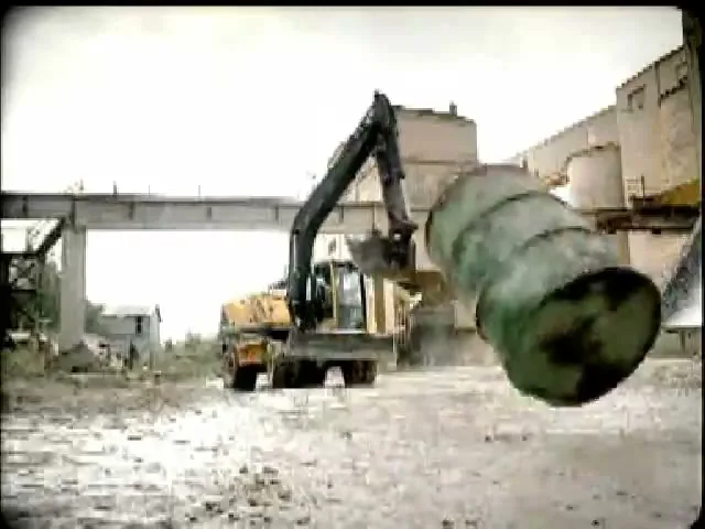 A yellow excavator is seen from a low angle, with a large green barrel falling towards the camera in a dusty industrial area.