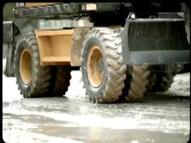 A close-up shot of a large, dirty construction vehicle's wheels on a wet, muddy surface, with water splashing as it moves.