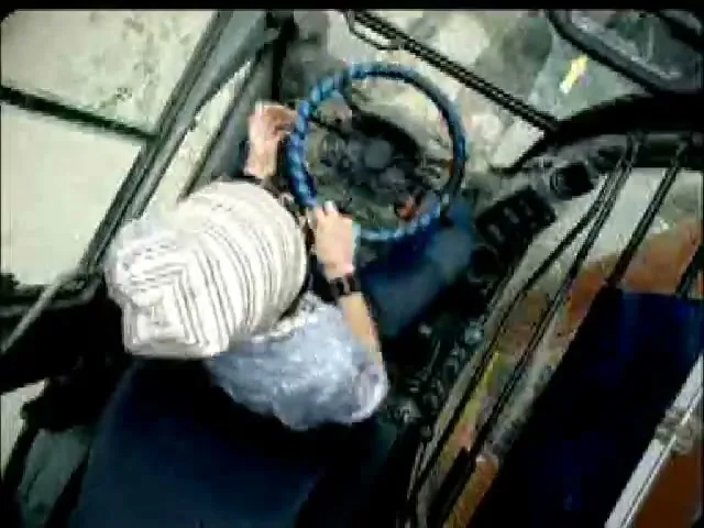 A man in a striped hat and blue shirt struggles to turn the steering wheel of a large vehicle, viewed from an overhead perspective inside the cab.