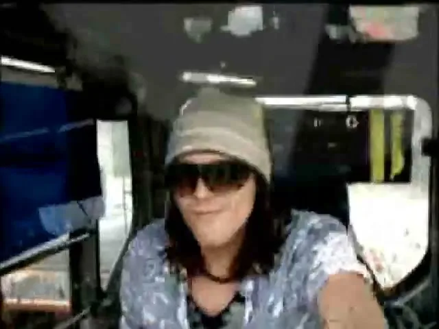A young man with long hair, a beanie, and sunglasses is seen from the chest up, looking at the camera while driving a vehicle during the day.
