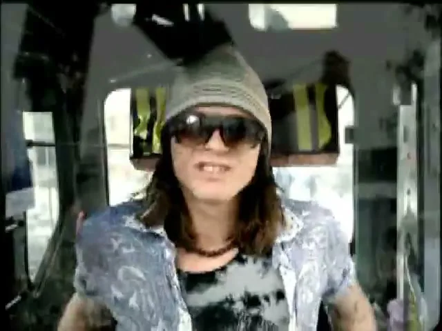 A young man with long hair, a beanie, and sunglasses is seen from the chest up, making a face inside a vehicle.