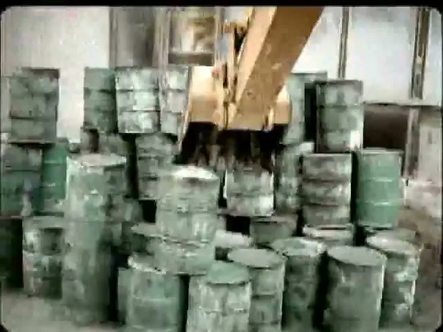 A yellow excavator arm with a claw picks up a green barrel from a large stack of barrels in an industrial setting.