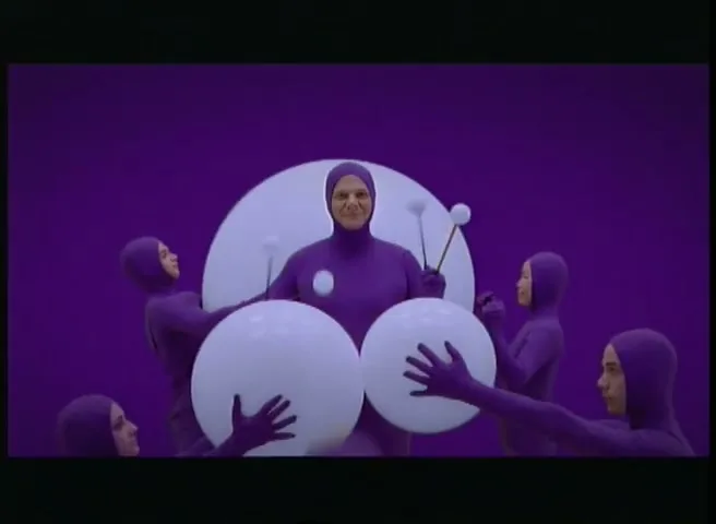 A man in a purple bodysuit juggles white balls as other people in purple bodysuits dance around him on a purple background.