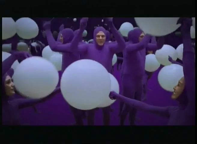 A crowd of people in purple bodysuits dance with white balloons in a purple void.