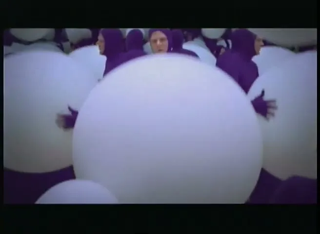 People in purple bodysuits push large white spheres around a purple floor in a studio.