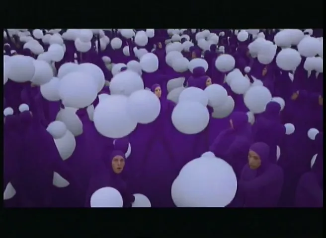 A crowd of people in purple bodysuits are surrounded by white balloons, some of which they are holding, in a surreal, abstract setting.