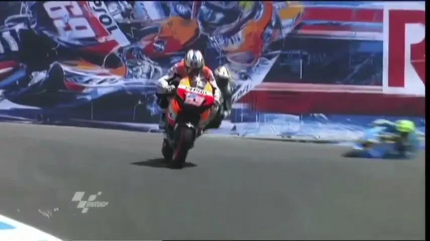 Two motorcycle racers in full gear speed around a track, with one racer falling behind the other, on a sunny day.