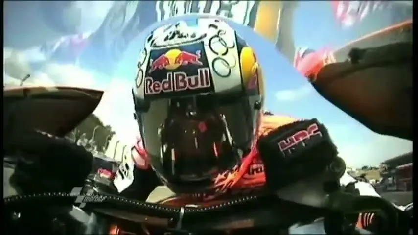 A motorcyclist in a helmet with a Red Bull logo is seen from a POV shot, racing on a track under a bright sky.