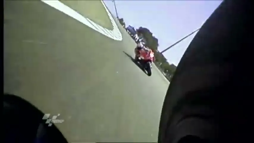 A motorcycle racer in a red and white suit leans into a turn on a race track, filmed from a low angle behind another motorcycle.