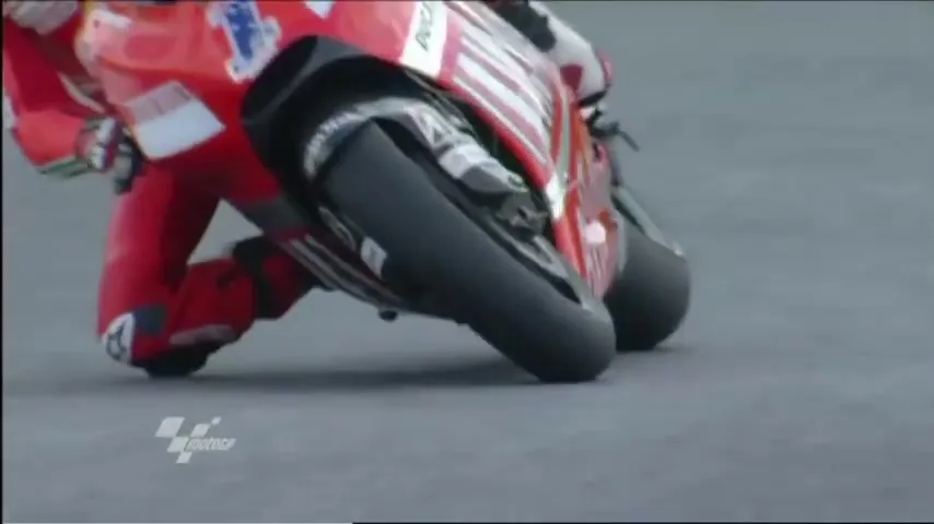 A male motorcycle racer in a red suit leans into a turn on a race track, with the camera focusing on the front wheel.