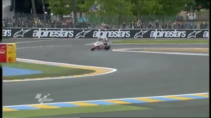 A motorcycle racer in white and black gear crashes into a yellow and red barrier on a race track, with spectators in the background.