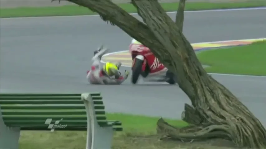 A motorcycle racer in a red and white suit crashes on a track, sliding across the asphalt near a tree and a green bench.