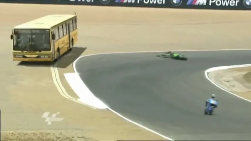 A yellow bus is parked on the side of a race track as a motorcyclist crashes into a green motorcycle on the track.
