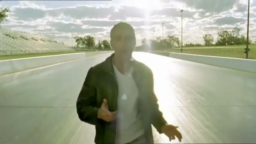 A man with gray hair and a dark jacket walks towards the camera on a racetrack, speaking directly to the viewer under a bright, cloudy sky.