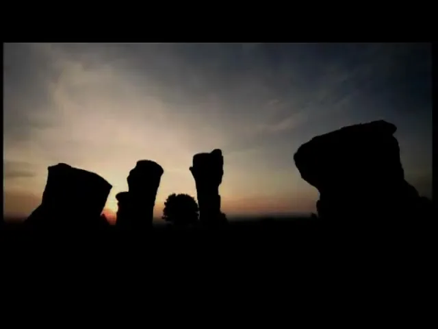 Silhouetted rock formations stand against a vibrant sunrise or sunset sky, with a small tree visible in the middle ground.