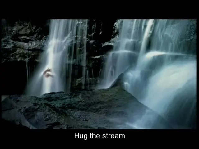 A person's arm reaches out from behind a powerful waterfall, with dark rocks and flowing water visible in a natural setting.
