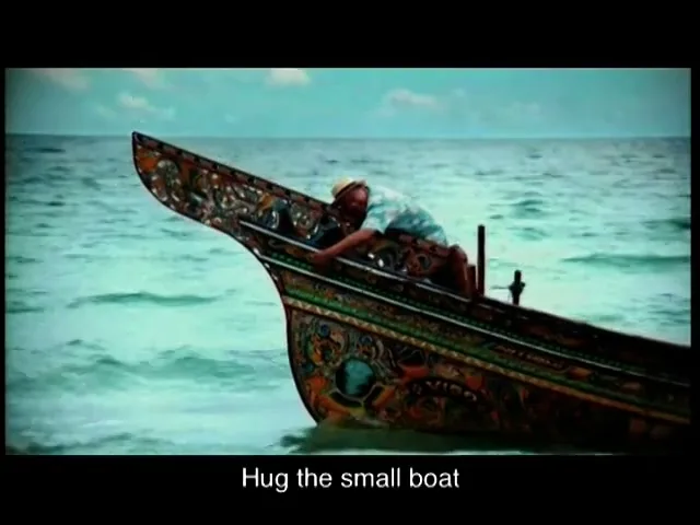 A man in a straw hat and light blue shirt hugs the bow of a small, ornately decorated wooden boat in the turquoise ocean under a blue sky.