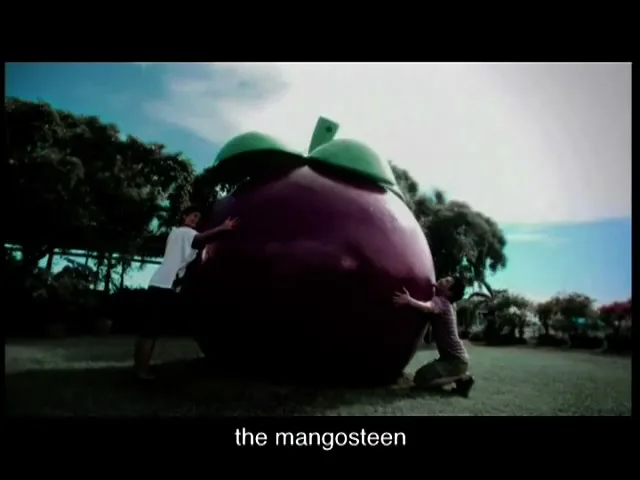 Two young men hug a giant mangosteen sculpture in a park under a bright blue sky, showing affection for the fruit.