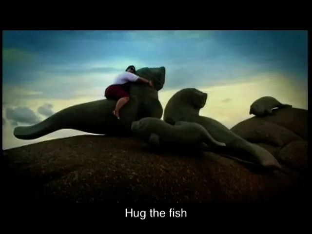 A man in a white shirt and red shorts hugs a large, dark grey dugong statue on a rocky outcrop under a cloudy sky.