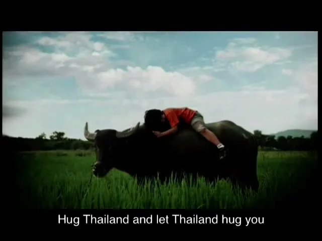 A young boy in a red shirt and shorts rides a water buffalo in a lush green field under a cloudy sky, then hugs its neck.