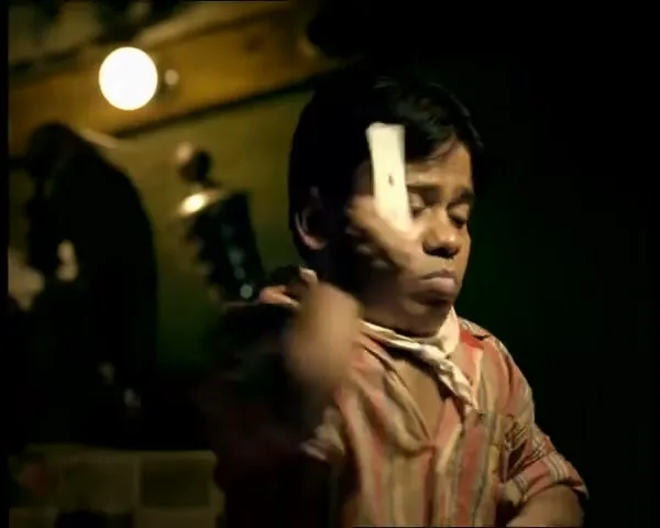 A man with dwarfism in a striped shirt and neckerchief, looking serious, rubs a playing card on his cheek in a dimly lit interior.