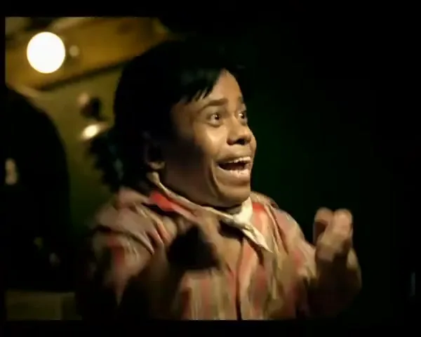 A man with short dark hair and a striped shirt reacts with wide eyes and an open mouth, raising his fists in excitement in a dimly lit interior.