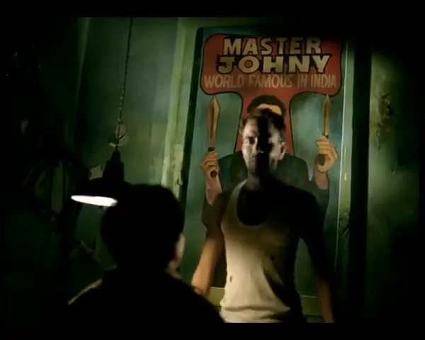 Two men are in a dimly lit room, one in the foreground with his back to the camera, and the other in a white tank top standing in front of a "Master Johnny" poster.