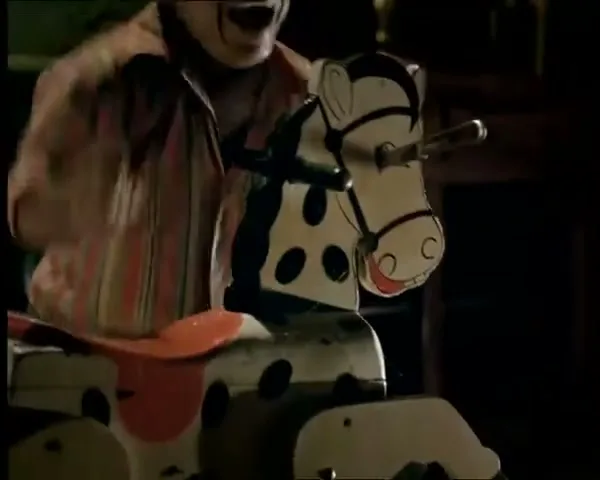 A man with a painted clown face and striped shirt rides a rocking horse in a dimly lit room, laughing maniacally.