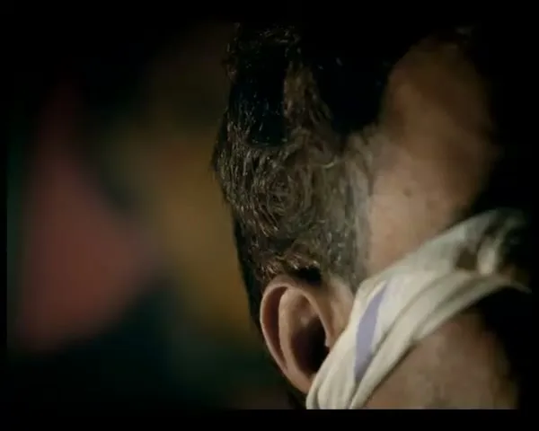 A man with dark, curly hair and a white cloth tied around his face is seen in a dark, interior setting.
