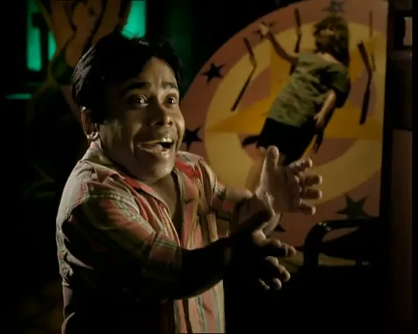 A man with a worried expression claps his hands in front of a spinning wheel with a woman tied to it, in a dark carnival setting.