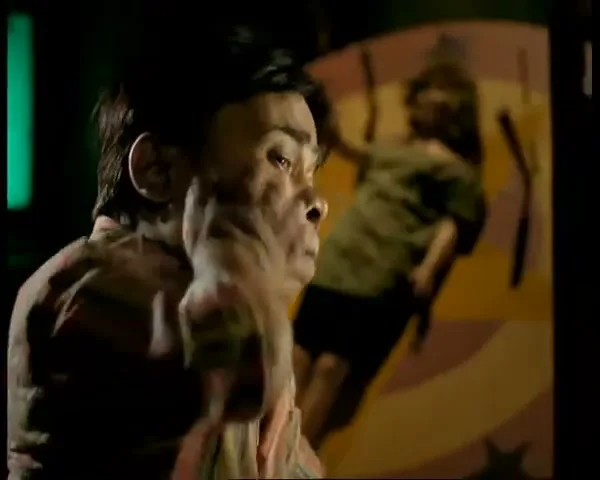 A man with dwarfism in a plaid shirt looks on as his face is distorted by an unseen force, in a dark carnival setting.