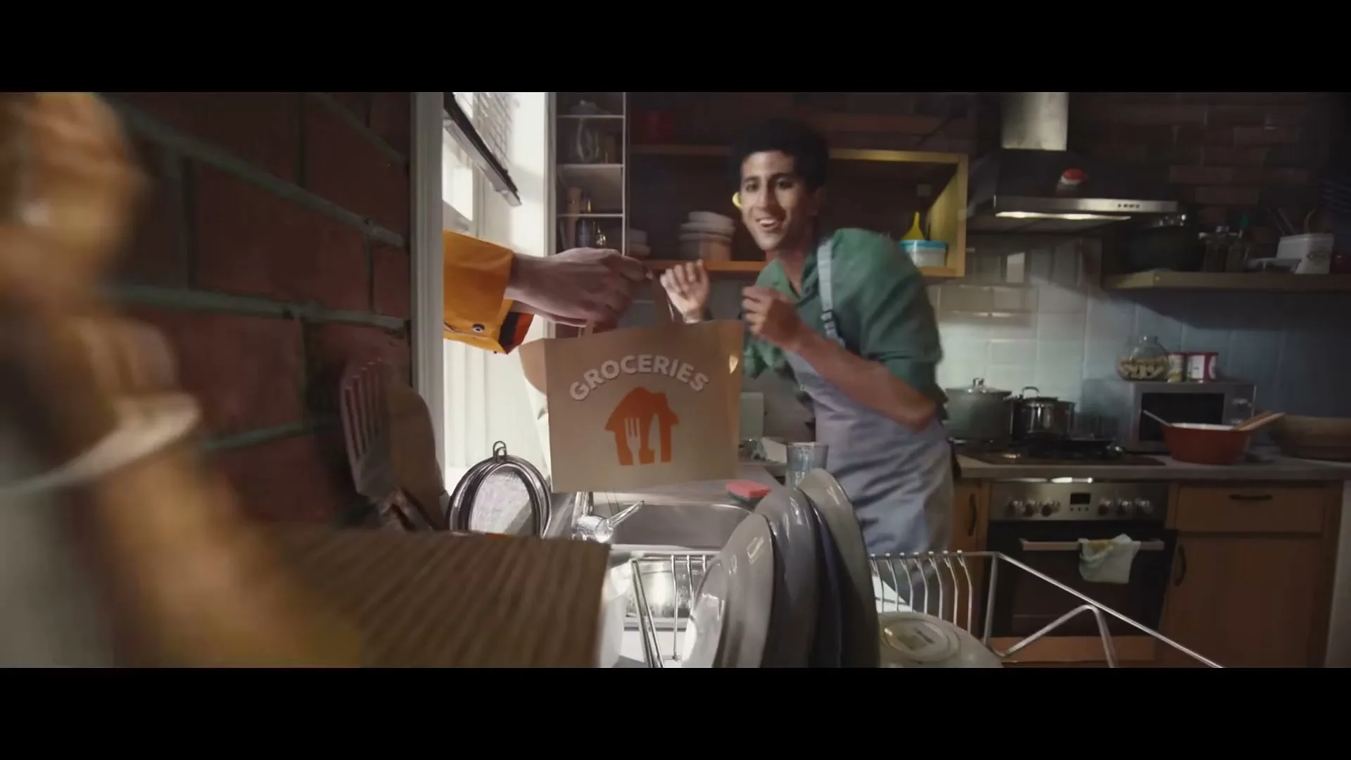 A man in an apron receives a grocery bag from an unseen person through a kitchen window, while a miniature version of a famous person on a timer talks on the phone.