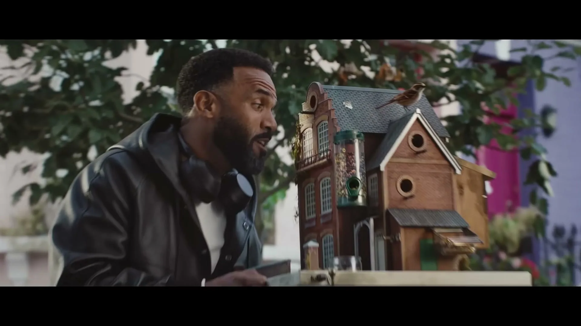 A man with a beard and headphones around his neck looks at a miniature birdhouse with a bird on top, outdoors.