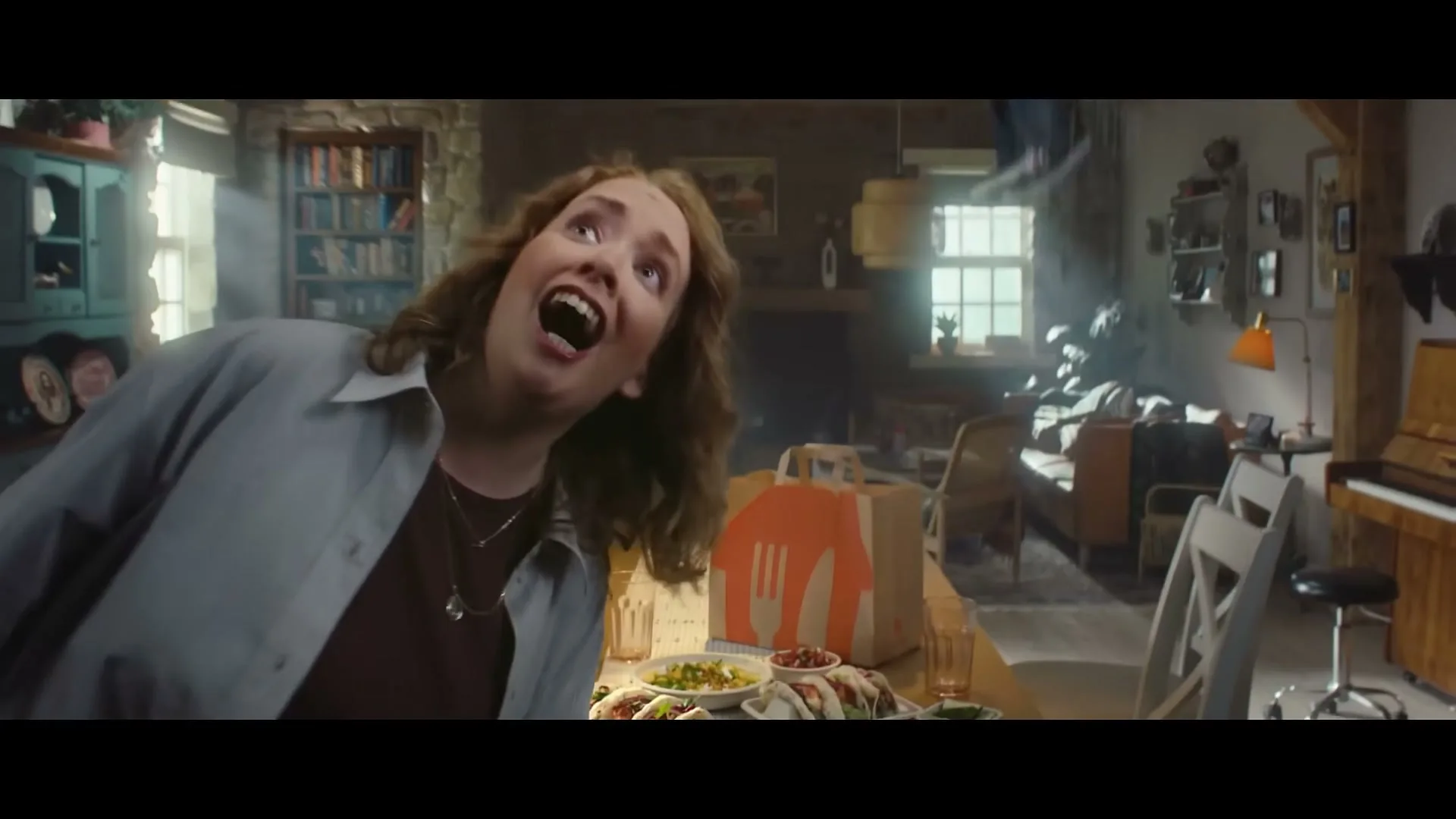 A woman with brown hair in a blue shirt and brown top looks up in surprise as a table full of tacos and corn is set in a rustic dining room.