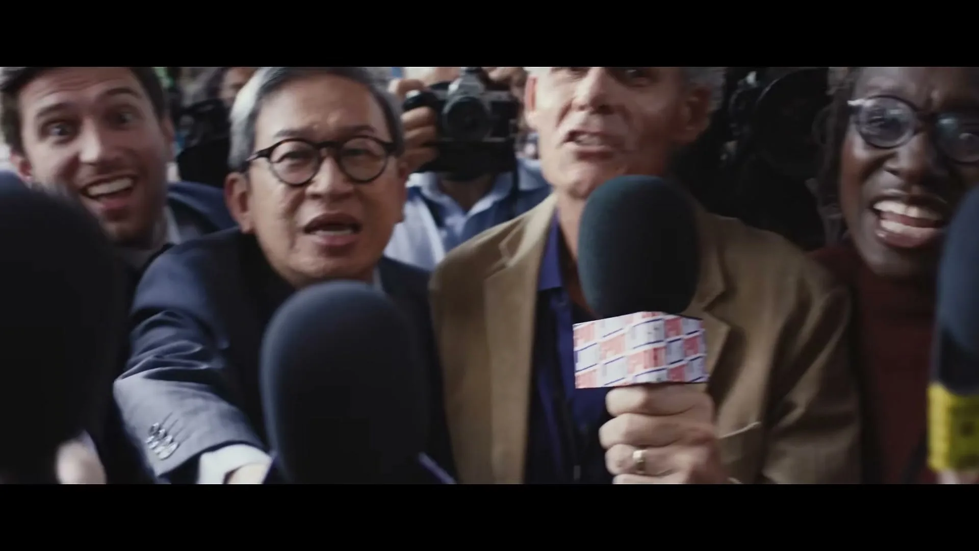 A diverse group of reporters, holding microphones, eagerly question a man in a brown jacket at an outdoor press conference.