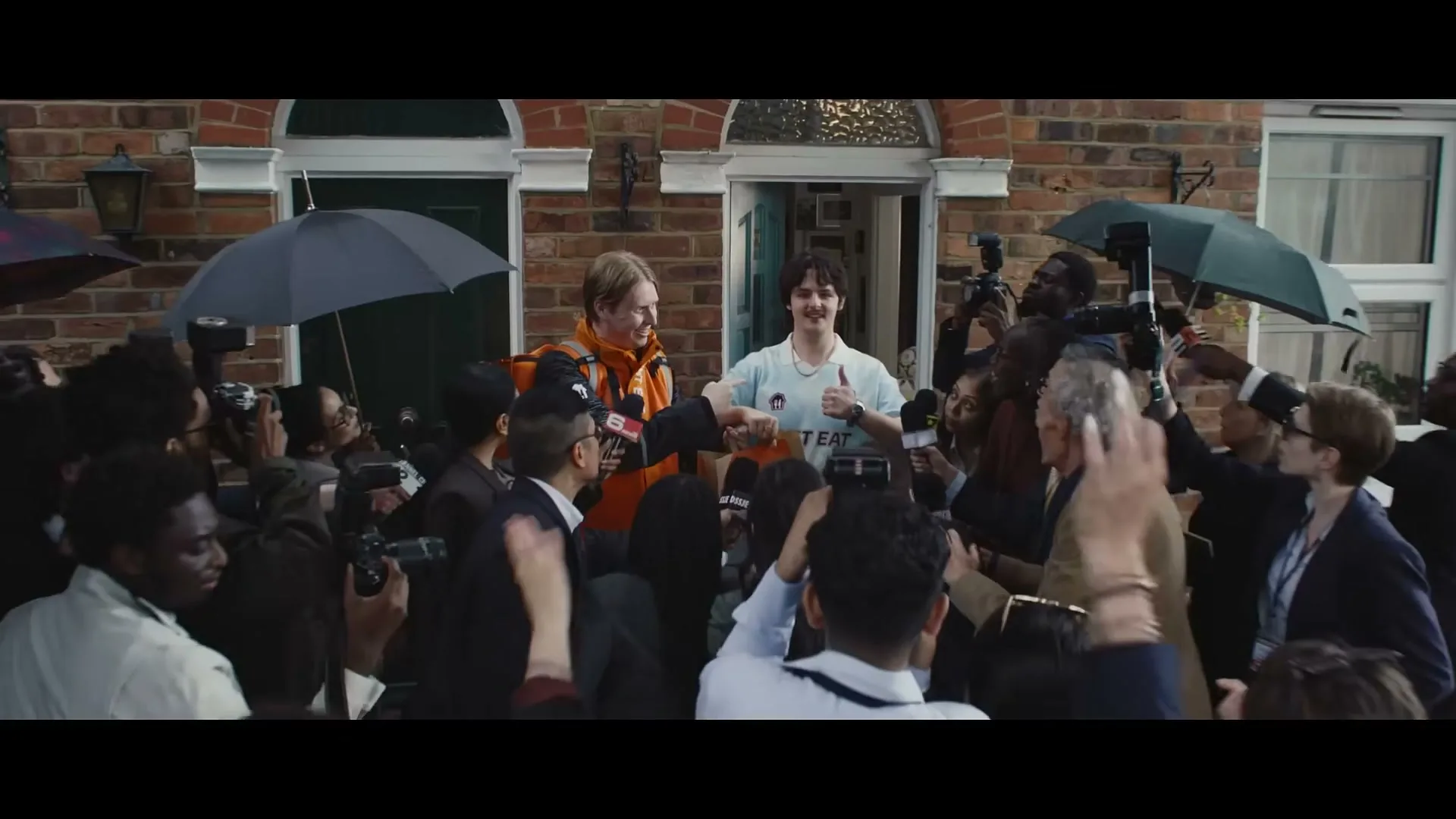A delivery driver in an orange jacket and a young man in a light blue shirt fist bump, surrounded by a crowd of paparazzi and reporters outside a brick building.