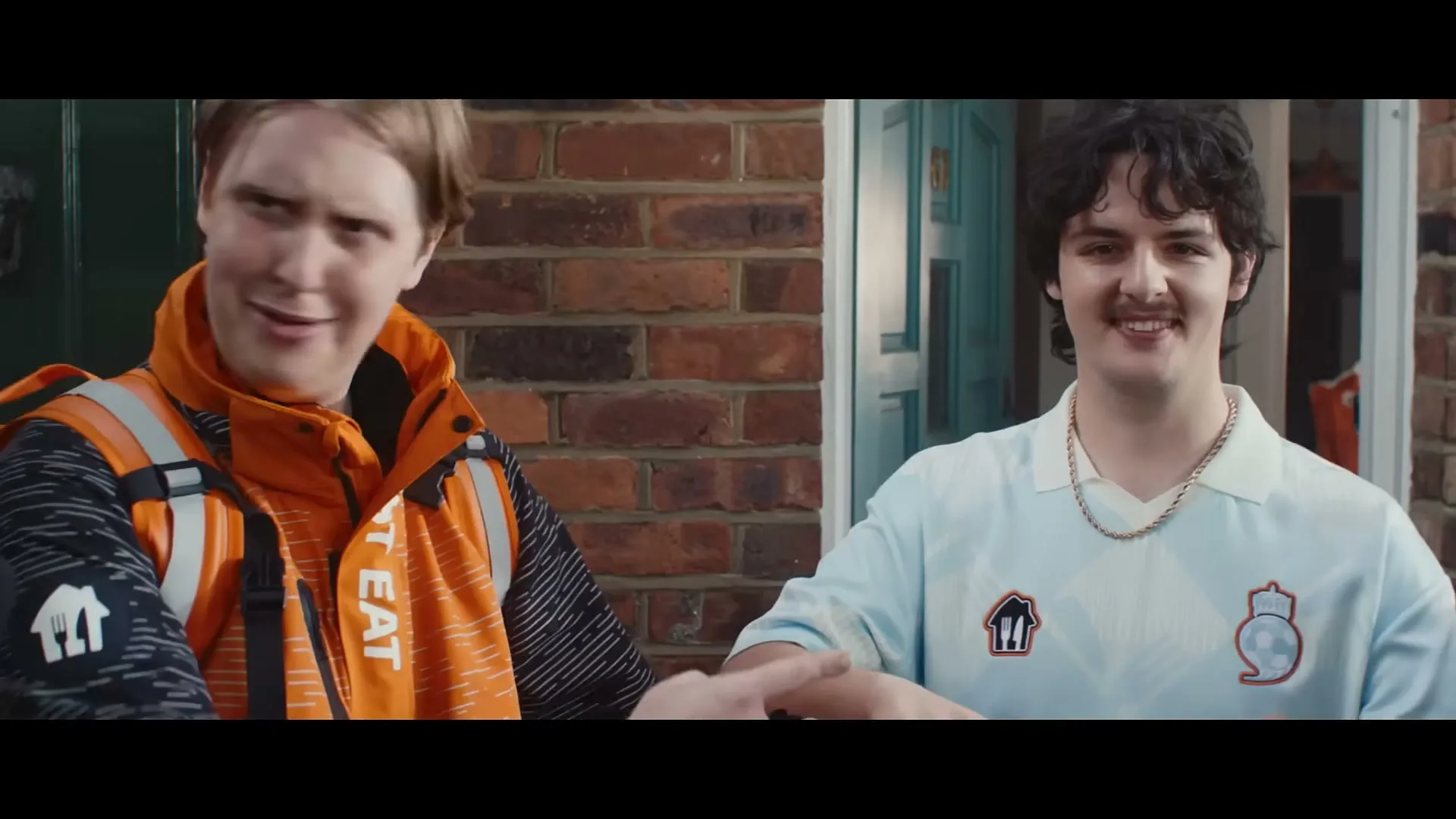 A delivery driver in an orange jacket and a man in a light blue jersey shake hands in front of a brick house.