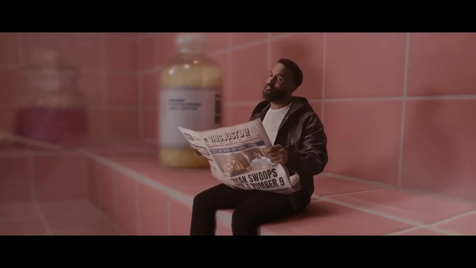 A man with a beard sits on a pink tiled bench, reading a newspaper in a pink tiled room, looking up and then down at the paper.