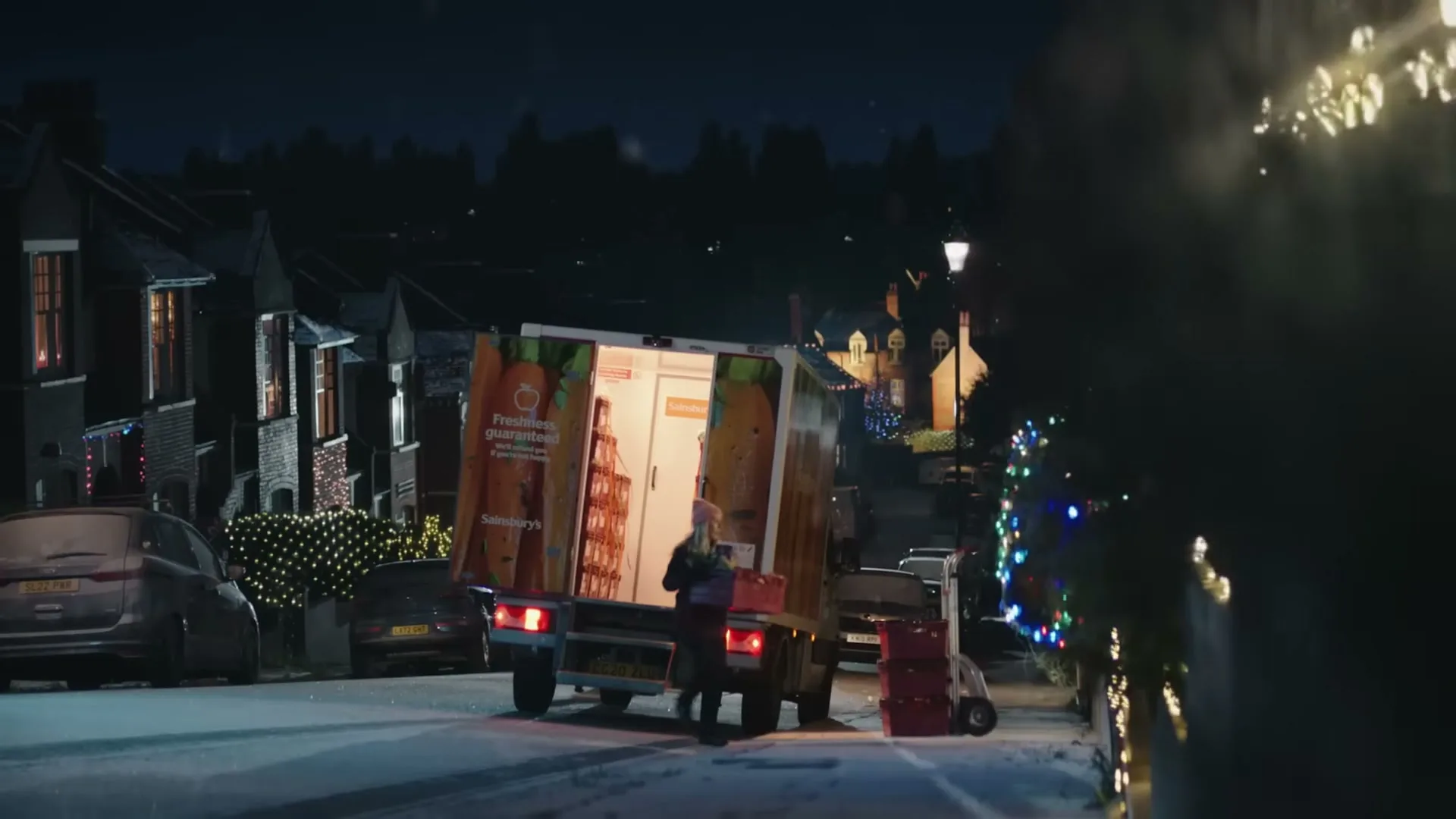 A delivery driver in a winter hat and coat unloads groceries from a Sainsbury's truck on a snowy, decorated street at night.