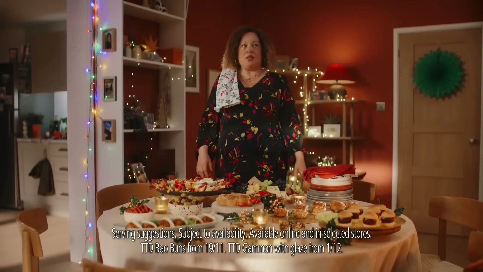 A woman in a floral dress with a towel over her shoulder stands behind a table laden with food, then walks away in a festive living room.