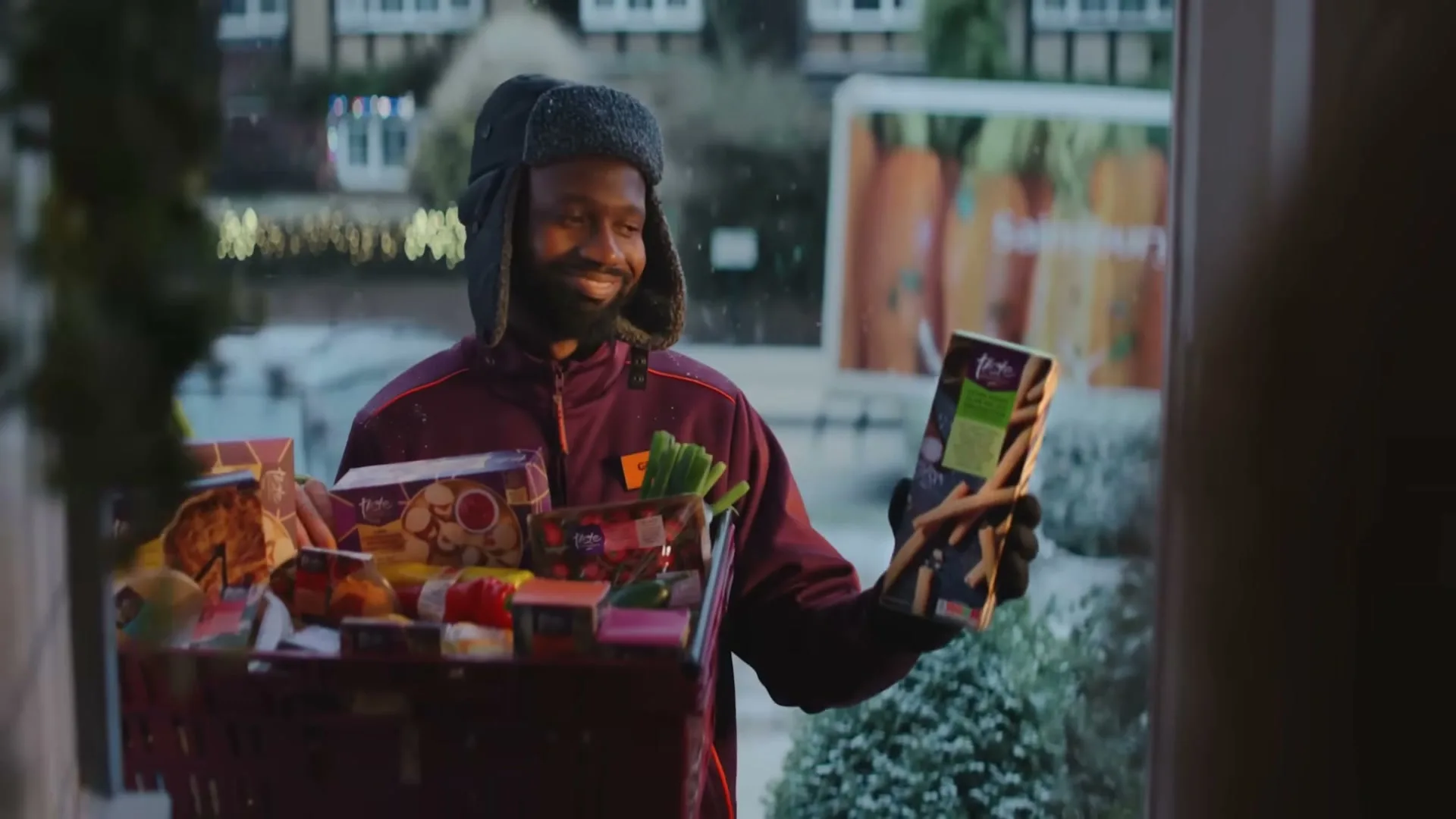 A delivery driver, a Black man in his 30s, smiles while holding a crate of groceries and a box of breadsticks at a snowy doorstep.