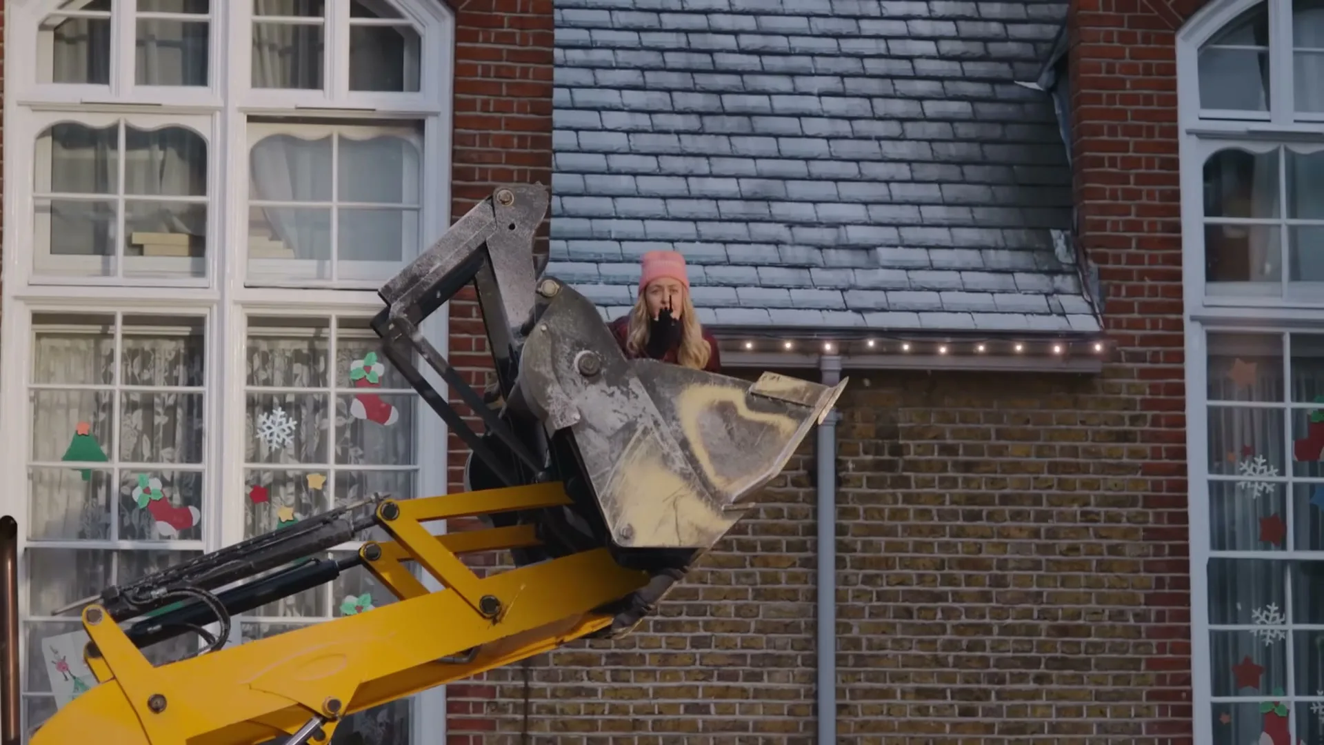 A young woman in a pink hat is lifted in the bucket of an excavator, looking through binoculars at a house decorated for Christmas.