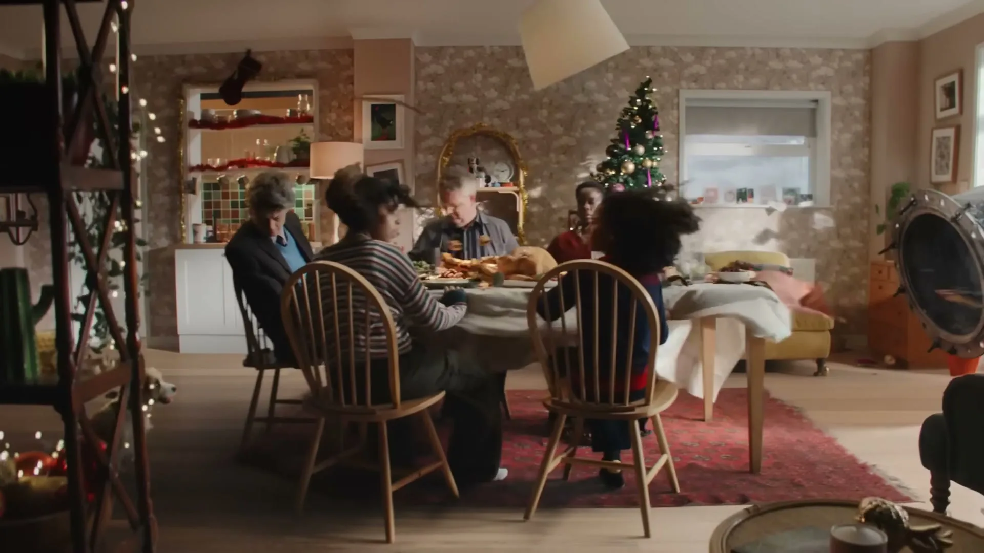 A diverse family of five, including a child, sits around a dining table laden with food, celebrating Christmas in a cozy, decorated home.