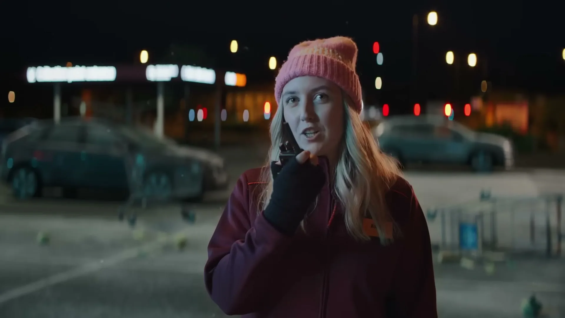 A young woman in a pink beanie and maroon jacket speaks into a walkie-talkie in a supermarket parking lot at night.