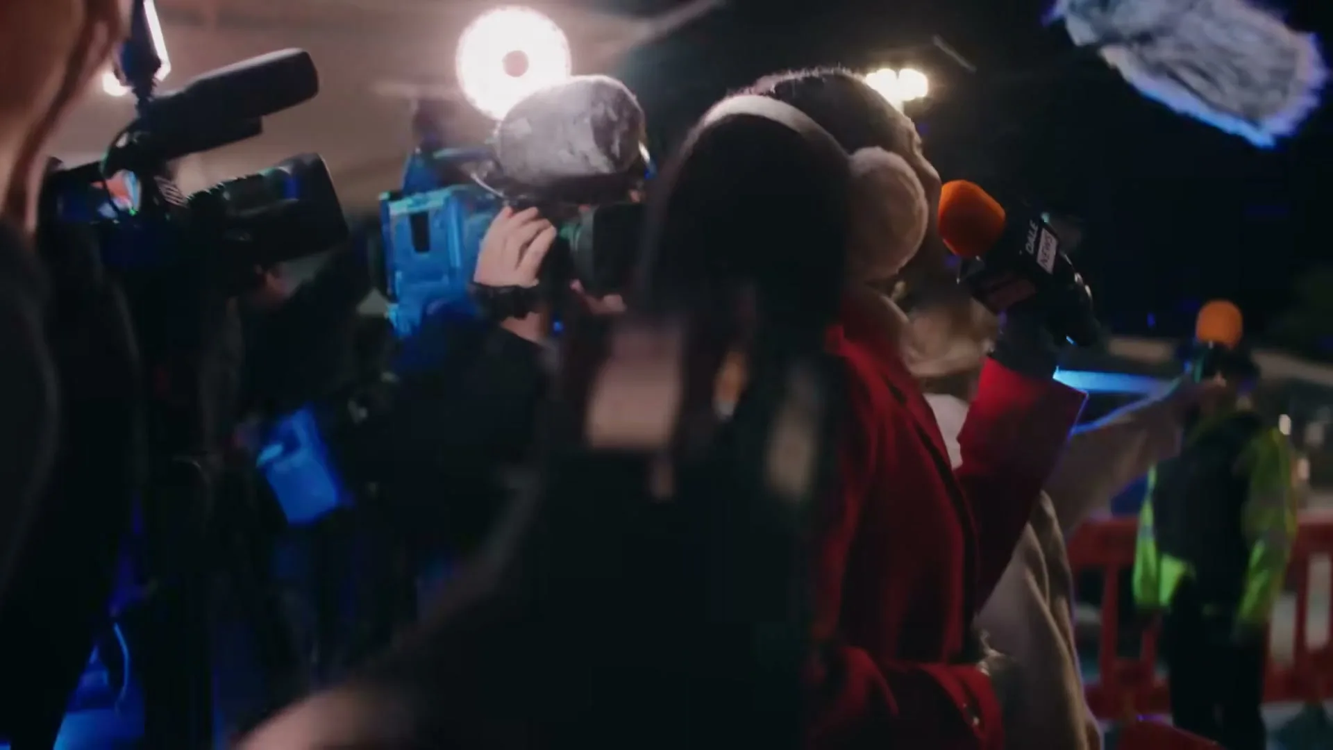 A female reporter in a red coat and earmuffs holds a microphone, surrounded by a crowd of paparazzi and camera operators at night.