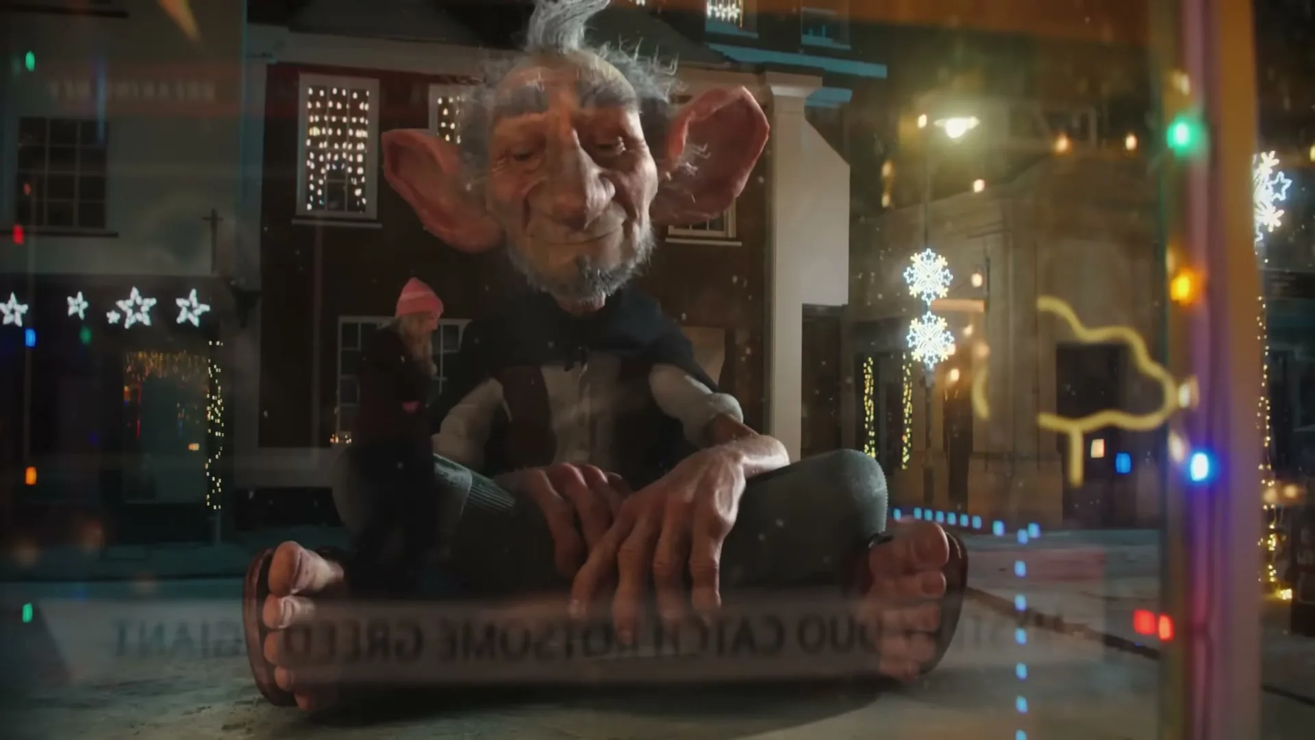 A giant-like creature with large ears and a beard sits cross-legged on a snowy street, looking at a small girl in a pink hat, surrounded by Christmas lights.