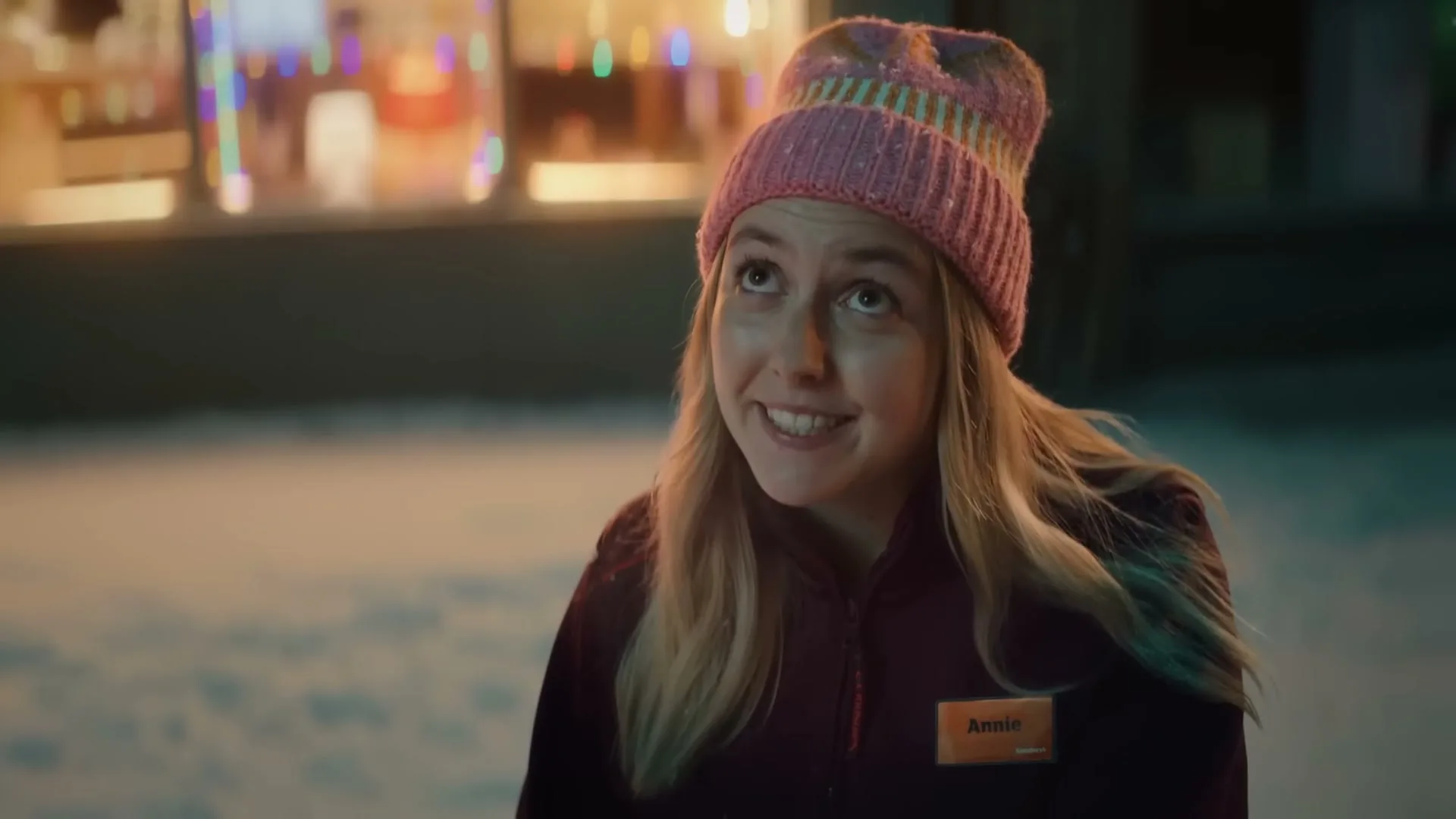 A young woman with blonde hair and a pink beanie looks up with a hopeful expression, standing in the snow outside a store decorated with Christmas lights.
