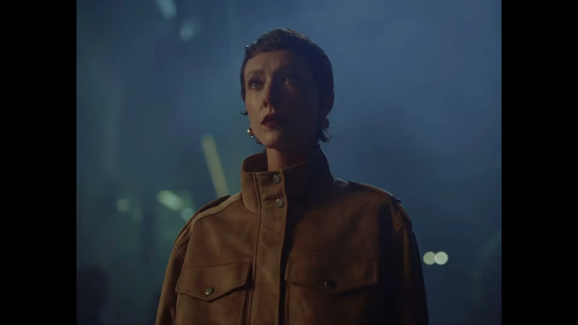 A woman in a brown jacket slowly lifts her head and looks up with a serious expression in a dark, smoky interior.